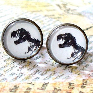 May include: A pair of bronze-colored earrings with a white background and a black silhouette of a dinosaur skeleton.