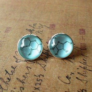 May include: Two round stud earrings with a silver border. Each earring has a light blue, transparent centre with a black chemical structure design. The earrings are presented on a brown, textured surface with handwritten text.