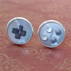 May include: A pair of silver-toned stud earrings featuring a video game controller design. One earring displays a black cross, while the other shows three gray and white buttons. The earrings are set against a patterned background.