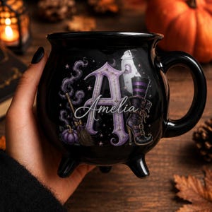 May include: A black cauldron-shaped ceramic mug featuring a large purple letter A with the name Amelia written in white script. The design includes a broomstick, a purple pumpkin, and a black and purple striped boot with stars and smoke.