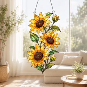 Op de afbeelding: Een glas-in-lood zonnevanger met drie grote gele zonnebloemen met donkerbruine harten en twee kleinere zonnebloemknoppen. Het bloemstuk bevat groene bladeren en stengels en hangt aan een dunne metalen ketting voor een hangende presentatie.