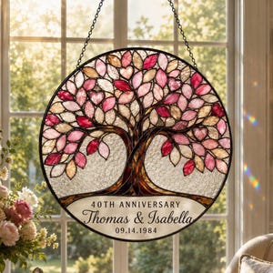 Op de afbeelding: Een ronde glas-in-lood zonnevanger met een boom met roze, rode en crèmekleurige bladeren. De basis bevat de tekst 40TH ANNIVERSARY, Thomas & Isabella en 09.14.1984. Het object hangt aan een zwarte metalen ketting voor een raam.