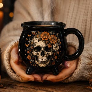 May include: A black ceramic cauldron-shaped mug featuring a detailed illustration of a human skull surrounded by orange and white roses and small bats. The mug has three short legs, a wide rim, and a curved handle, with steam rising from the top.