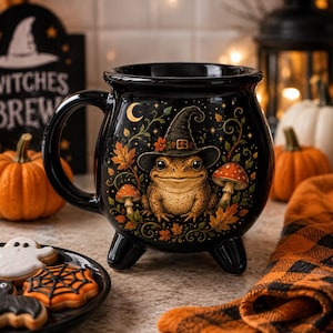 May include: A black ceramic cauldron-shaped mug featuring a graphic of a toad wearing a witch hat, surrounded by autumn leaves, mushrooms, and a crescent moon. The mug has three small feet and a curved handle, with a glossy finish and detailed art.
