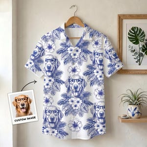 Puede incluir: Una camisa de manga corta con botones que presenta un patrón repetitivo en azul y blanco de perros golden retriever con gafas negras, intercalados con flores de hibisco tropicales y hojas de palma. Una pequeña impresión cuadrada muestra la imagen del perro.