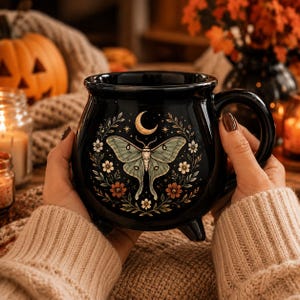 May include: A black ceramic cauldron-shaped mug featuring a central illustration of a pale green moth with outstretched wings. Above the moth is a golden crescent moon, surrounded by delicate floral vines with small white and orange flowers.