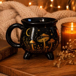 May include: A black ceramic cauldron-shaped mug featuring a gold-toned illustration of mushrooms, a crescent moon, and scattered stars. The mug has a rounded body, a curved handle, and three small feet at the base, with a glossy finish throughout.