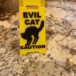 May include: A yellow caution sign with the words "EVIL CAT" above a black silhouette of a cat and the word "CAUTION" below. The sign is made of plastic and has a folding design.
