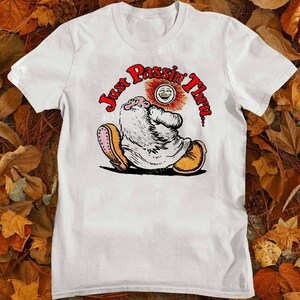 May include: White t-shirt featuring a whimsical illustration of a bearded character with the text "Just Passin' Thru" in red. The character is wearing yellow shoes and is seated with a smiling sun above them. The shirt is made of a soft material.