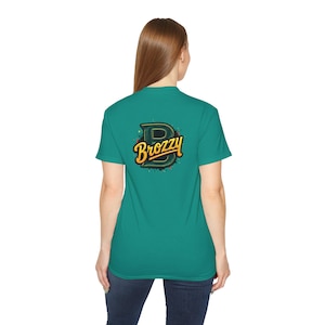 Può includere: Una maglietta color teal con la scritta "Brozzy" in un font stilizzato sul retro. La parola è gialla con un contorno nero e un design a spruzzi verdi e neri.
