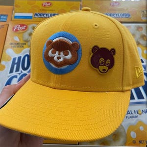 May include: A yellow baseball cap with a curved brim. The cap features two bear-shaped patches: one embroidered bear on a blue circle and a smaller, solid bear. The New Era logo is visible on the side.