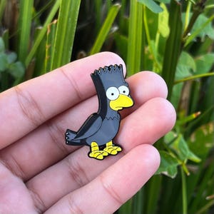 May include: A black and gray enamel pin in the shape of a cartoon bird with yellow feet and beak. The bird has a spiky black head and large white eyes. The pin is held in a hand against a background of green foliage.