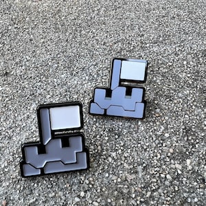 Könnte beinhalten: Zwei pixelige Emaille-Anstecknadeln. Jede Anstecknadel hat ein grau-weißes Design mit schwarzem Rand. Die Anstecknadeln haben eine flaggenartige Form und ein geometrisches Design. Die Anstecknadeln werden auf einer strukturierten grauen Oberfläche präsentiert.