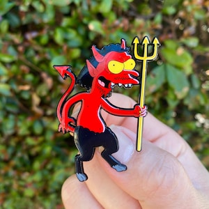 May include: A red devil enamel pin with black trousers and hair, yellow eyes, and a yellow trident. The devil has a red tail and is holding the trident in its right hand. The pin is set against a blurred green background.