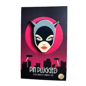 May include: A black and gray enamel pin featuring a stylized Catwoman mask design. The pin is set against a red circle and a black background with a cityscape silhouette. The text "PIN PLUGGED" is displayed below the pin.