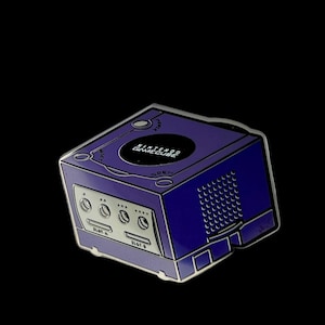 May include: A purple Nintendo GameCube pin with silver accents. The pin features the console's design, including the logo, buttons, and slots. The text "Nintendo GameCube" is visible on the top. The pin is set against a black background.