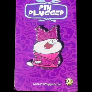 May include: A purple enamel pin featuring a cartoon character with a white body, pink hat, and pink outfit. The pin is on a purple backing card with the text "PIN PLUGGED" and a website address. The background has a swirling pattern.