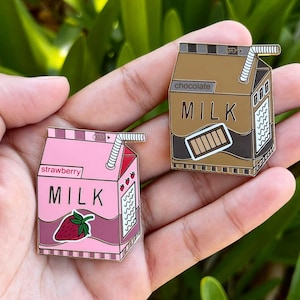 May include: Two enamel pins shaped like milk cartons. One is pink with "strawberry MILK" and a strawberry graphic. The other is brown with "chocolate MILK" and a chocolate bar graphic. Both have a straw.