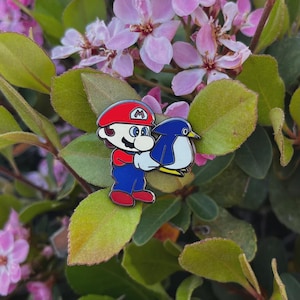 May include: A colorful enamel pin featuring a cartoon character in a red hat and blue overalls holding a blue and white penguin. The pin has a silver border and is set against a background of green leaves and pink flowers.