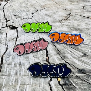 Może przedstawiać: Cztery kolorowe emaliowane przypinki z napisem "DOOM" w stylu graffiti. Przypinki są w kolorach zielonym, pomarańczowym, różowym i granatowym, każda z białym obrysem. Leżą na zwietrzałej drewnianej powierzchni.