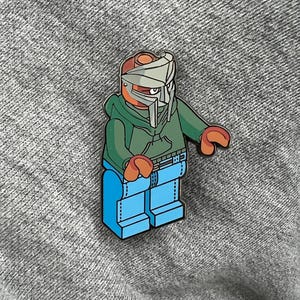 May include: A colourful pin featuring a stylized figure in a green hoodie, blue jeans, and a silver mask. The figure has orange hands and a red head. The pin is designed in a Lego-like style and is set against a grey background.