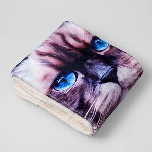 Peut inclure: Couverture pliée avec un gros plan du visage d'un chat aux yeux bleus vifs. La couverture a une texture douce et moelleuse avec un dessous de couleur crème. La fourrure du chat est un mélange de marron, de violet et de blanc.