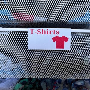 May include: A white metal mesh storage basket featuring a rectangular label with the red text T-Shirts and a red t-shirt icon. The basket contains various folded garments, and another identical mesh basket is visible above it.