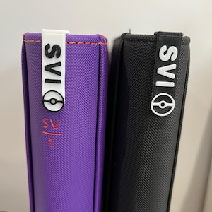 Puede incluir: Se muestran dos carpetas, una morada y otra negra, una al lado de la otra. La carpeta morada tiene costuras naranjas y texto rojo. Ambas carpetas tienen una etiqueta blanca con el texto "SVIAS" y un logotipo circular.