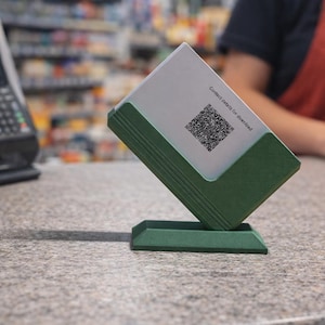 Puede incluir: Un tarjetero verde con una tarjeta blanca con un código QR y el texto "Contact details for download". El soporte está inclinado sobre una encimera cerca de un sistema de punto de venta.