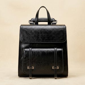 May include: Black leather backpack with a flap closure and a front pocket secured by two straps. The backpack has a top handle and adjustable shoulder straps. The leather has a slightly distressed appearance, giving it a vintage look. The hardware is a dark, gunmetal color.