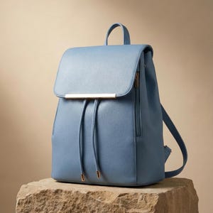 Può includere: Uno zaino azzurro con chiusura a patta e una barra color oro. Lo zaino ha una chiusura con coulisse e una piccola maniglia in alto. La borsa è appoggiata su una superficie di pietra.