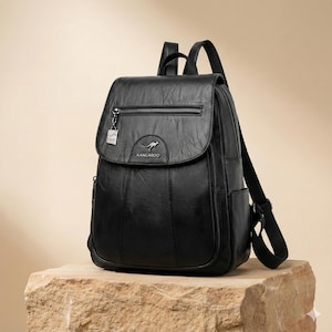 May include: Black leather backpack with a flap closure and a zippered front pocket. The backpack features a small metal tag and a kangaroo logo. The straps are adjustable. The bag is sitting on a stone surface.