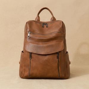 May include: A brown faux leather backpack with multiple zippered compartments and a top handle. The backpack has a large front pocket and two side pockets. The zippers are dark brown. The bag is designed for everyday use.