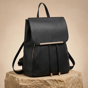 Può includere: Zaino nero con chiusura a patta e barra color oro. Lo zaino ha un manico superiore e spallacci regolabili. È dotato di chiusura con coulisse e una piccola tasca con cerniera laterale. Lo zaino è appoggiato su una pietra.