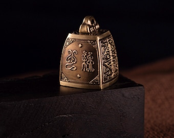 May include: A small, antique-style brass bell with intricate engraved patterns and Chinese characters. The bell is rectangular with a loop at the top for hanging. It sits on a dark wooden block against a black background.