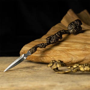May include: A metal letter opener featuring a sharp, pointed silver-toned blade and a dark, textured handle shaped like a gnarled branch with intricate, rounded, bronze-colored decorative elements along its length and at the end.
