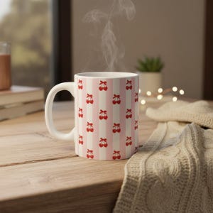 Könnte beinhalten: Weiße Keramiktasse mit einem rosa-weiß gestreiften Muster, verziert mit roten Kirschillustrationen. Die Tasse hat einen gebogenen Henkel und enthält ein dampfendes Getränk. Sie steht auf einer Holzoberfläche.