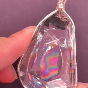 May include: A clear, faceted, teardrop-shaped crystal pendant encased in silver-colored wire wrapping. The interior of the crystal displays a vibrant, multi-colored rainbow refraction pattern. The letter B is visible in the bottom right corner.
