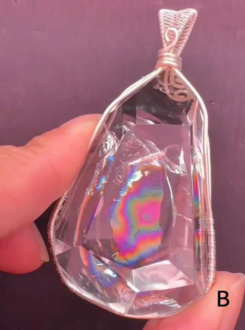 May include: A clear, faceted, teardrop-shaped crystal pendant encased in silver-colored wire wrapping. The interior of the crystal displays a vibrant, multi-colored rainbow refraction pattern. The letter B is visible in the bottom right corner.