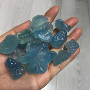 Puede incluir: Primer plano de una mano sosteniendo varias piedras preciosas de aguamarina en bruto. Las piedras, de color azul claro, presentan formas irregulares y una textura natural. La imagen destaca el aspecto bruto y auténtico de estas gemas.