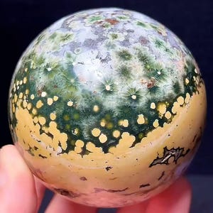 May include: A polished spherical stone featuring a complex pattern of deep forest green starburst inclusions, creamy yellow circular spots, and translucent white areas. The surface displays a smooth, glossy texture with natural mineral variations.