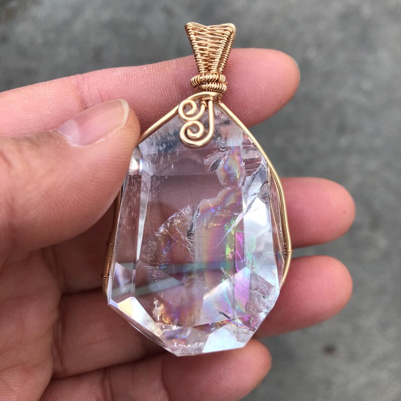 May include: A clear, faceted crystal pendant encased in a gold-colored wire wrap. The crystal features internal fractures that create a rainbow-like iridescent effect. The wire is coiled at the top to form a bail for hanging the pendant.