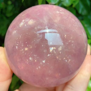May include: A polished, translucent rose quartz sphere. The sphere is a soft pink color with subtle internal inclusions and a reflective surface. The background is blurred green foliage.