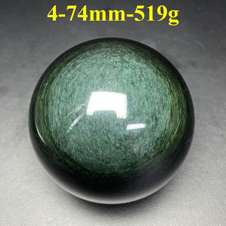 May include: A polished, dark green obsidian sphere. The surface reflects light, revealing a swirling pattern within the stone. The text "4-74mm-519g" is in the upper left corner. The sphere is approximately 2.9 inches in diameter and weighs 18.3 ounces.