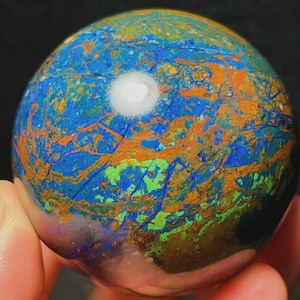 Puede incluir: Una piedra esférica pulida que presenta una mezcla vibrante de vetas azul profundo, verde brillante y naranja terroso. La superficie muestra un patrón marmoleado complejo con un acabado suave y brillante y un reflejo de luz blanco en la parte superior.
