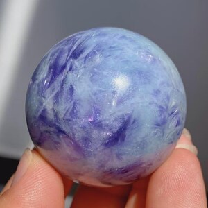 May include: A polished, spherical gemstone with a swirling pattern of light blue and deep purple hues. The crystal ball is held in a person's hand, showcasing its smooth, reflective surface. The gemstone is a decorative item.