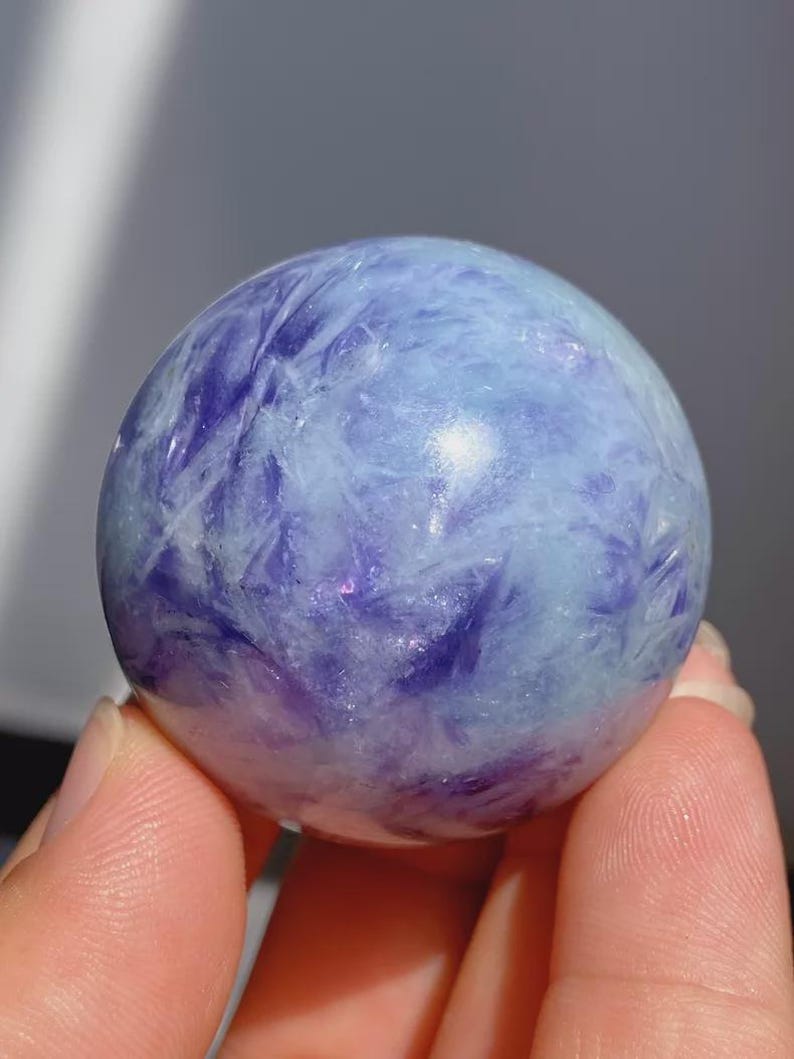 May include: A polished, spherical gemstone with a swirling pattern of light blue and deep purple hues. The crystal ball is held in a person's hand, showcasing its smooth, reflective surface. The gemstone is a decorative item.
