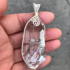 May include: A clear, faceted quartz crystal pendant encased in a silver-colored wire wrap. The wire features a decorative spiral design at the top, forming a bail for suspension. The crystal has visible internal fractures and natural inclusions.