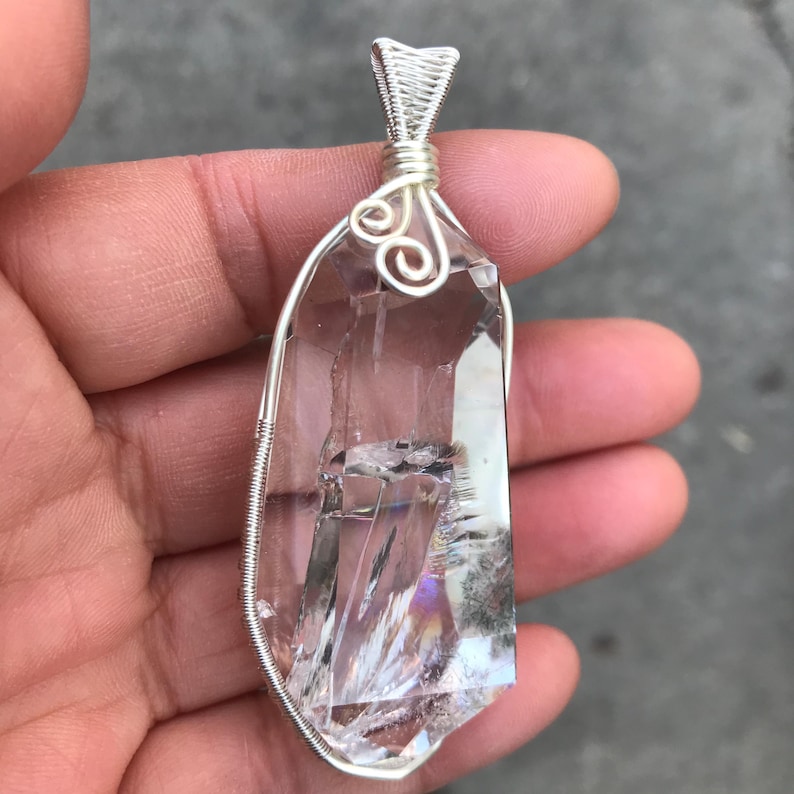 May include: A clear, faceted quartz crystal pendant encased in a silver-colored wire wrap. The wire features a decorative spiral design at the top, forming a bail for suspension. The crystal has visible internal fractures and natural inclusions.