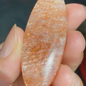Op de afbeelding: Een ovaalvormige zonnesteen cabochon met een doorschijnende, warme oranje tint en opvallende gouden, glinsterende insluitingen over het hele oppervlak. De gepolijste edelsteen heeft een gladde, reflecterende textuur.
